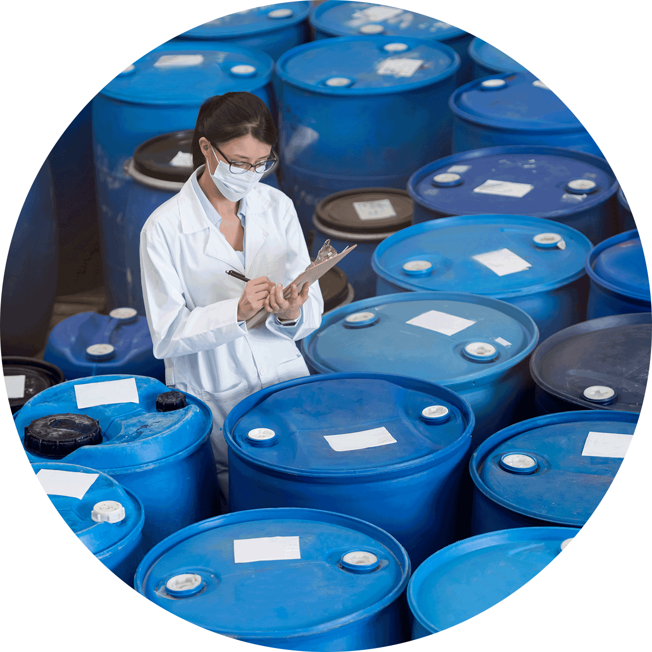 A quality control specialist in a white lab coat and face mask inspecting and documenting chemical storage barrels in a warehouse environment.